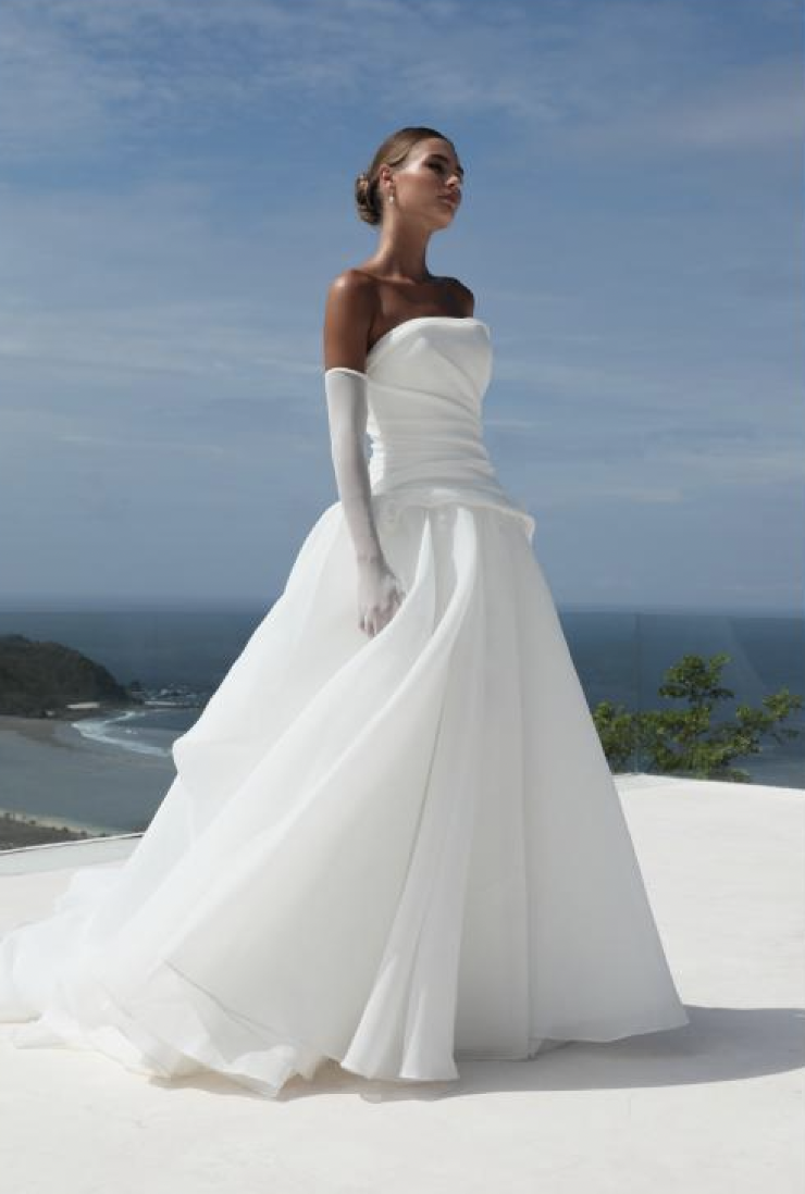 A woman in a strapless white wedding dress stands outdoors near a coastline, with an ocean view in the background. She has her hair styled elegantly and wears long white gloves.