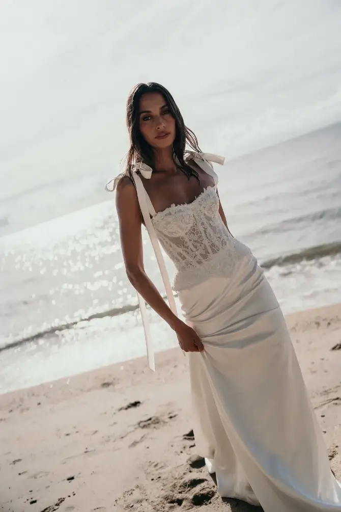 A woman wearing a white dress stands on a sandy beach with the ocean in the background.