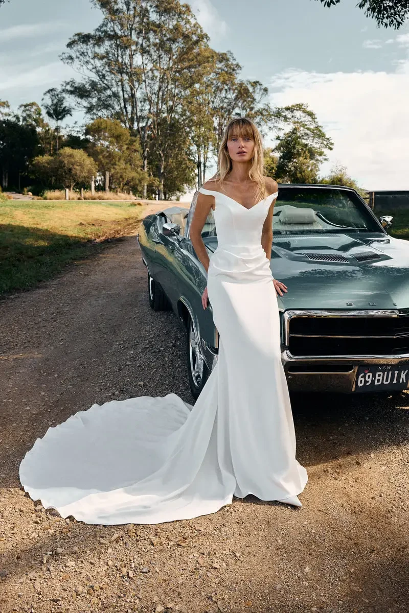Model is wearing a satin off-the-shoulder gown with side-ruched waist detail and a long, flowing train for a timeless silhouette.