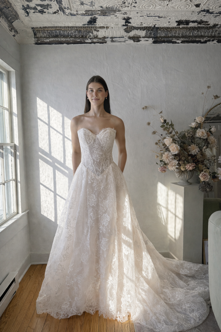 A model wearing a strapless white lace wedding gown stands in a well-lit room with large windows, showcasing the dress's intricate details. There are flowers in a vase nearby.