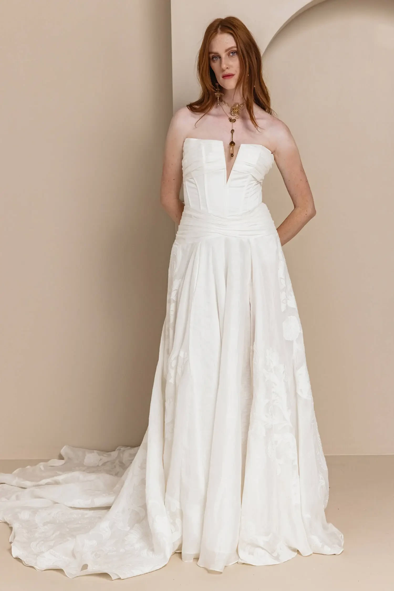 Model is wearing a corset gown with a soft v-neckline, drop waist, and embroidered linen skirt with deep folds and a full train.