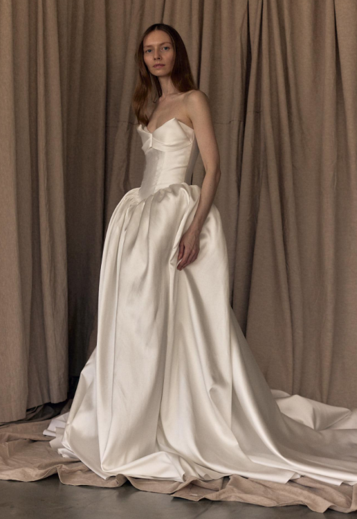 A model wearing an elegant white strapless gown with a fitted bodice and voluminous skirt, posed in front of a draped fabric background.