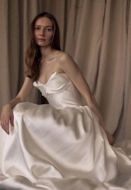A model wearing an elegant white strapless gown with a fitted bodice and voluminous skirt, posed in front of a draped fabric background.