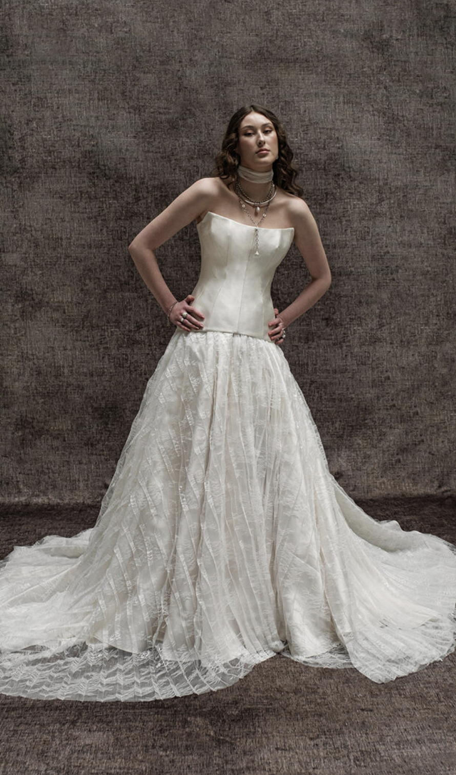 A model wearing a strapless white wedding gown with a full skirt, standing confidently with her hands on her hips against a textured background.