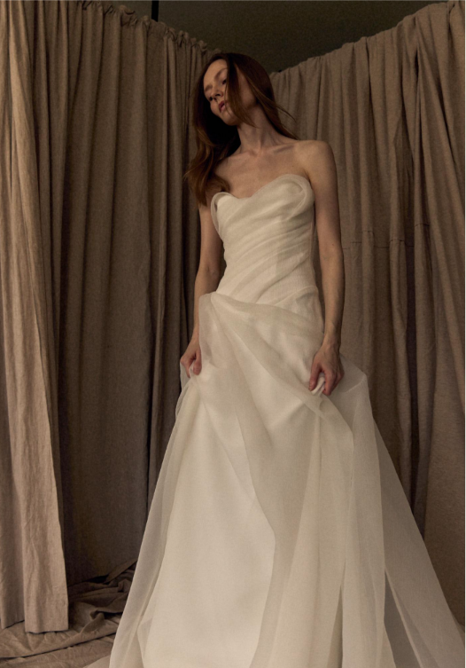 A woman in a strapless, flowing white gown standing against a backdrop of draped fabric.