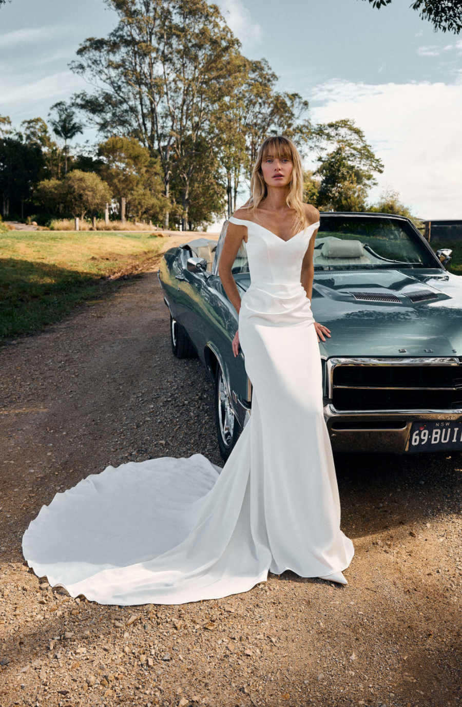 Model is wearing a luxurious satin off-the-shoulder gown with delicate ruching at the waist, a statuesque silhouette, and a long, elegant train.