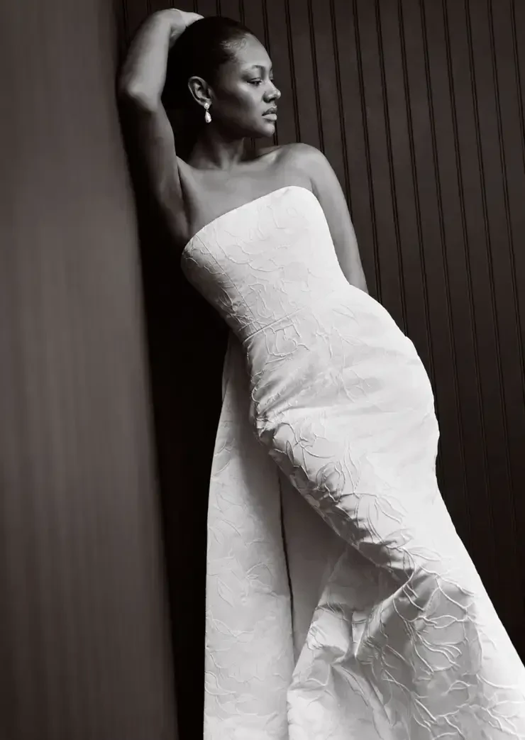 A woman in a strapless white dress leaning against a wall, captured in black and white.