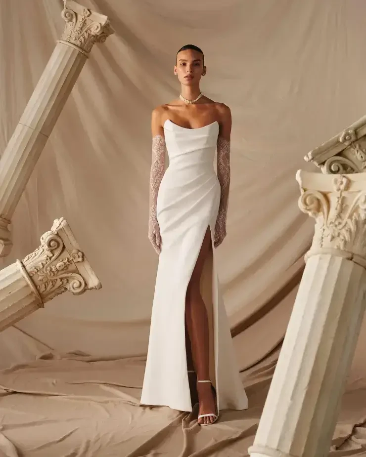 A model wearing a white off-shoulder gown with a slit, standing in front of a neutral backdrop with decorated columns.