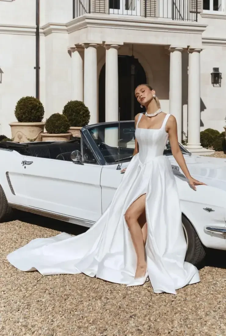 A woman in a long white dress with a high slit stands beside a white convertible car in front of a luxurious building.