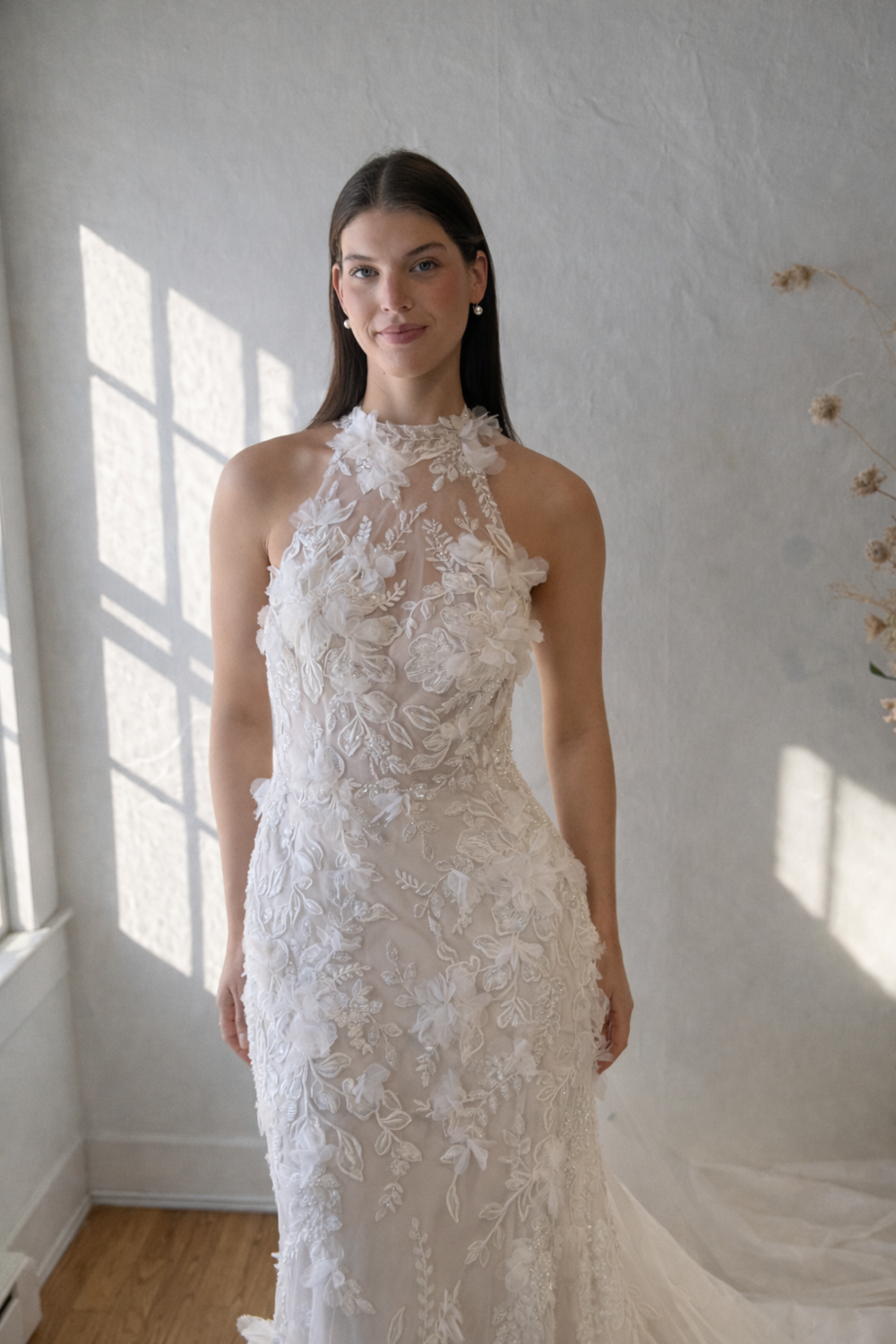A woman wearing a beautifully detailed, white wedding dress with floral embroidery, standing in a well-lit room.