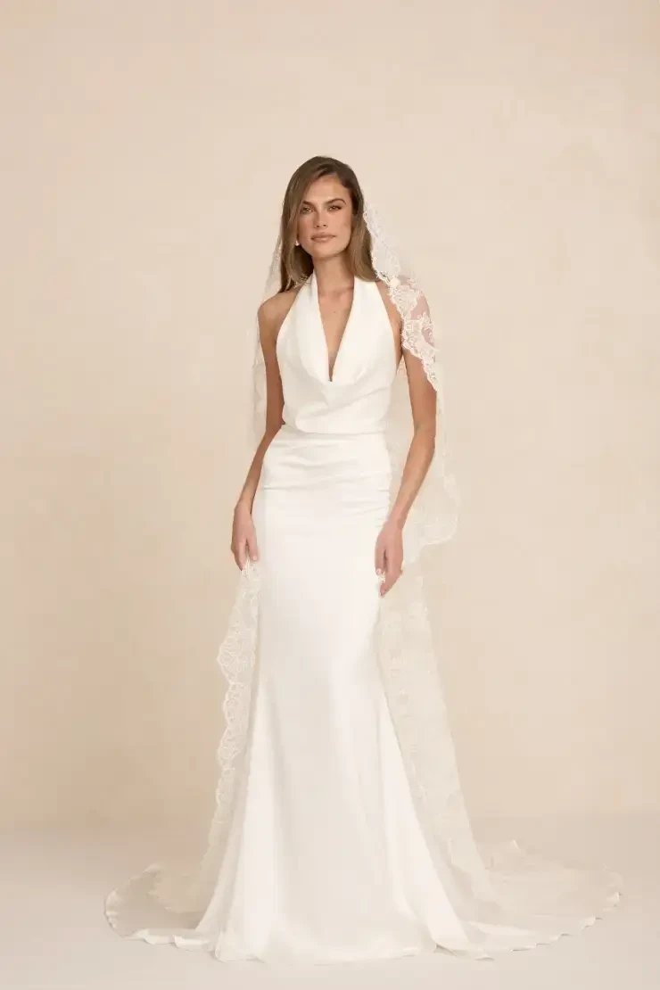A model wearing a sleek, elegant white wedding gown with a halter neckline and a sheer lace shawl.