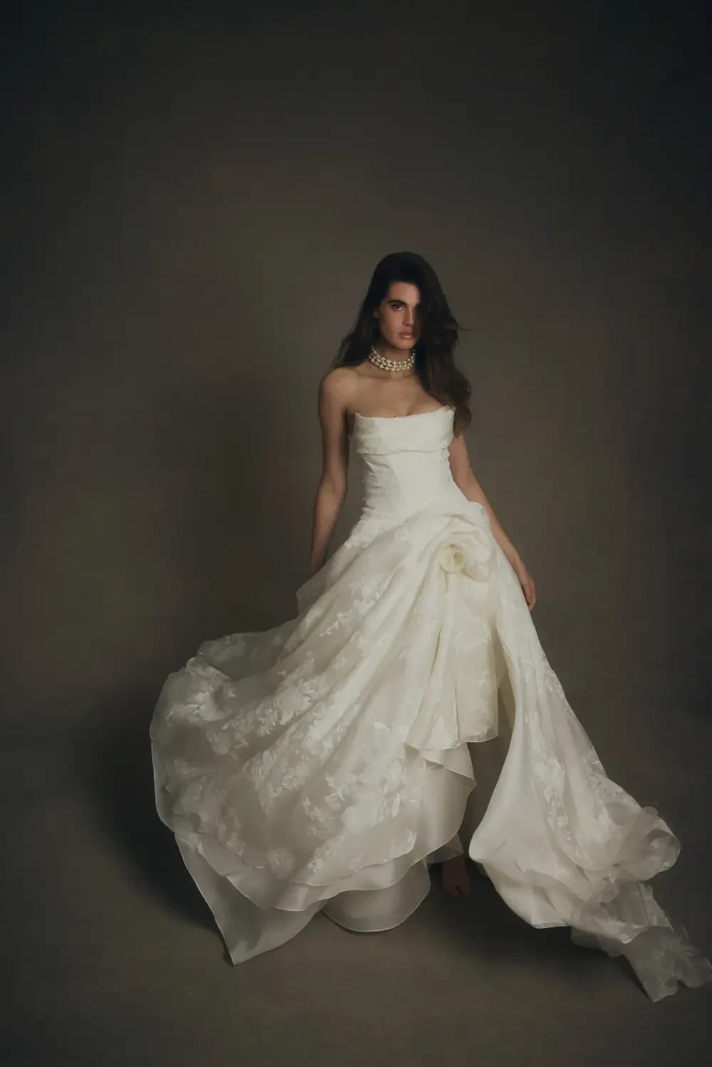 A woman in a strapless white gown with a dramatic flowing skirt, standing against a dark backdrop.